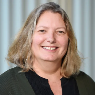A smiling woman with long brown hair wearing a black top and a pearl necklace stands in front of a blurred background.