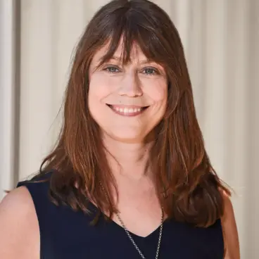A woman with long brown hair smiles at the camera. She is wearing a sleeveless navy blue top and a necklace with a pendant. The background is a light-colored curtain.