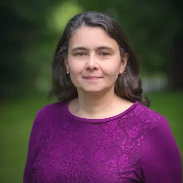 A woman with long dark hair is standing outdoors, wearing a purple top. She is looking at the camera with a neutral expression. The background is a blurred green, suggesting a natural setting.
