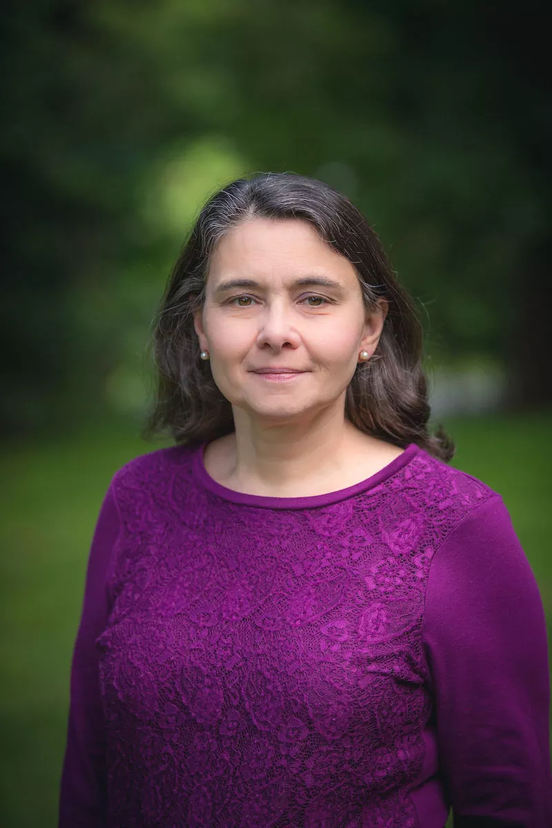 A woman with long dark hair is standing outdoors, wearing a purple top. She is looking at the camera with a neutral expression. The background is a blurred green, suggesting a natural setting.