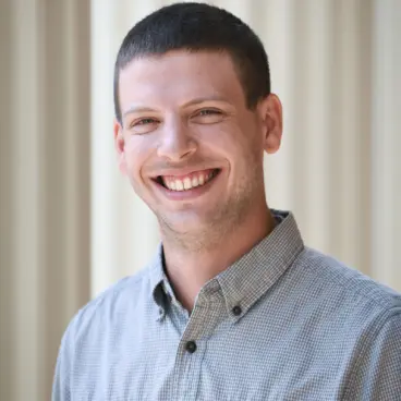 A person with short dark hair is smiling while wearing a gray button-up shirt. They are standing in front of a light-colored, blurred background.