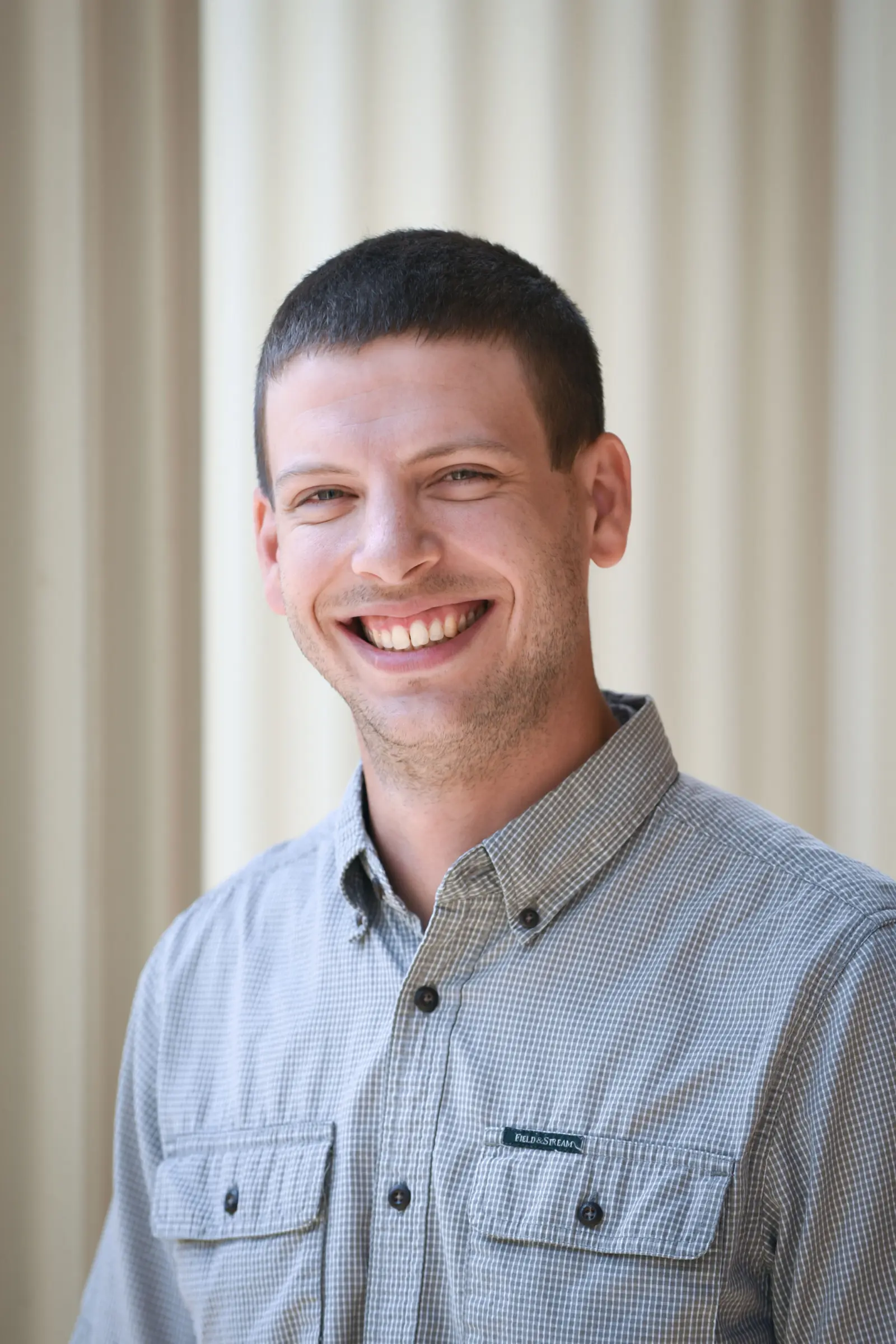 A person with short dark hair is smiling while wearing a gray button-up shirt. They are standing in front of a light-colored, blurred background.