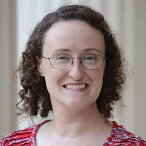 A person with curly brown hair, wearing glasses and a red patterned top, smiling in front of a blurred light-colored background.