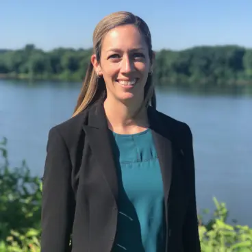 A woman with long hair smiles, wearing a black blazer and teal top. She is standing outdoors in front of a body of water with trees in the background under a clear blue sky.