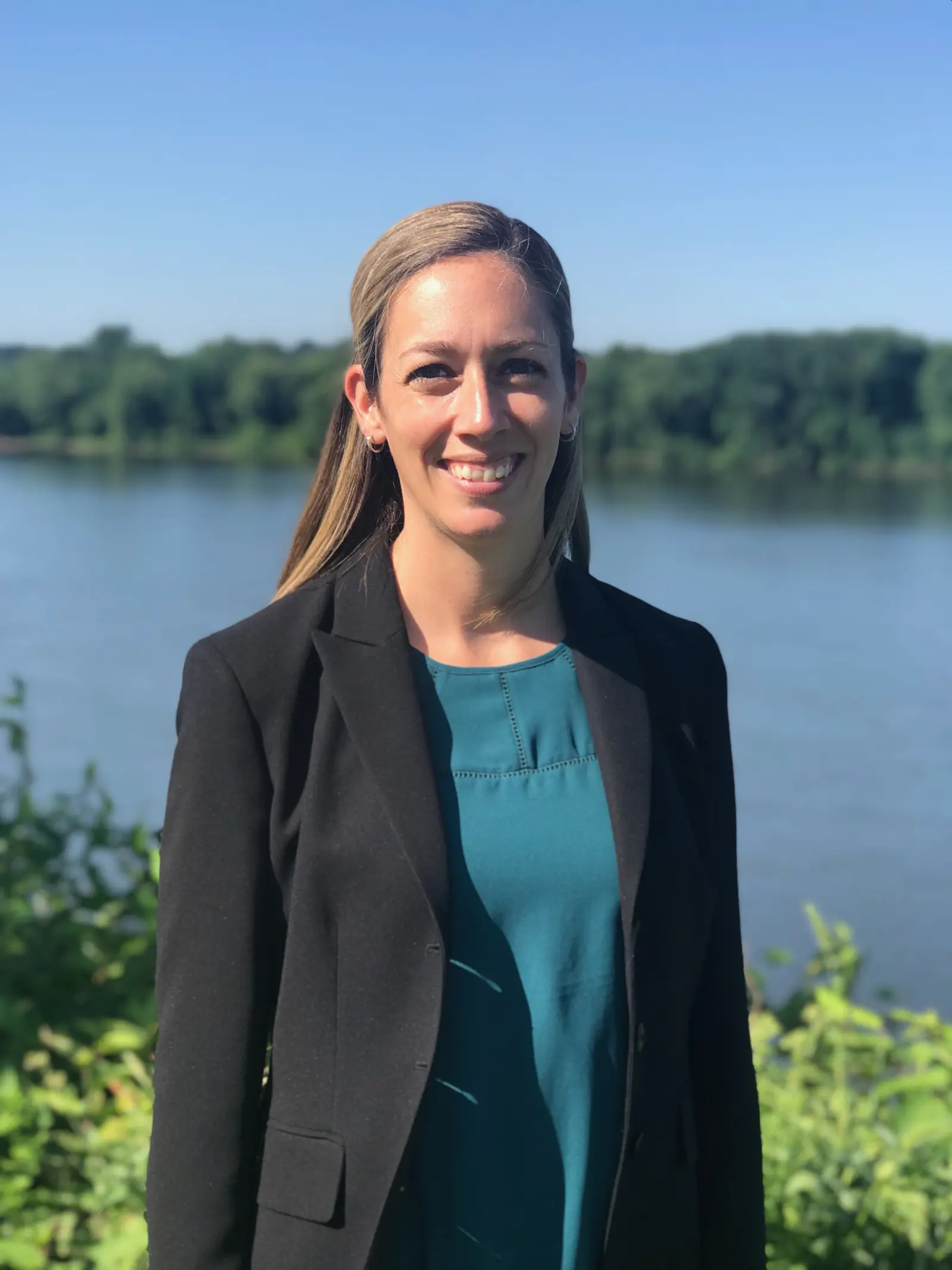 A woman with long hair smiles, wearing a black blazer and teal top. She is standing outdoors in front of a body of water with trees in the background under a clear blue sky.