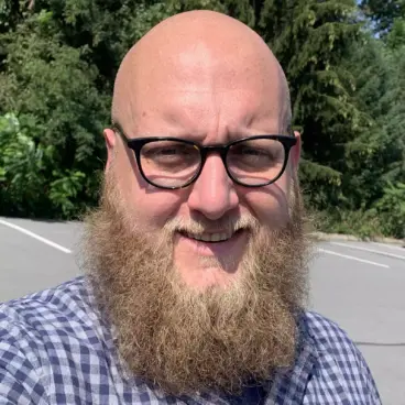 A smiling person with a bald head, glasses, and a full beard is wearing a checkered shirt. They are outdoors, standing in a parking area with trees in the background and a sunny sky.