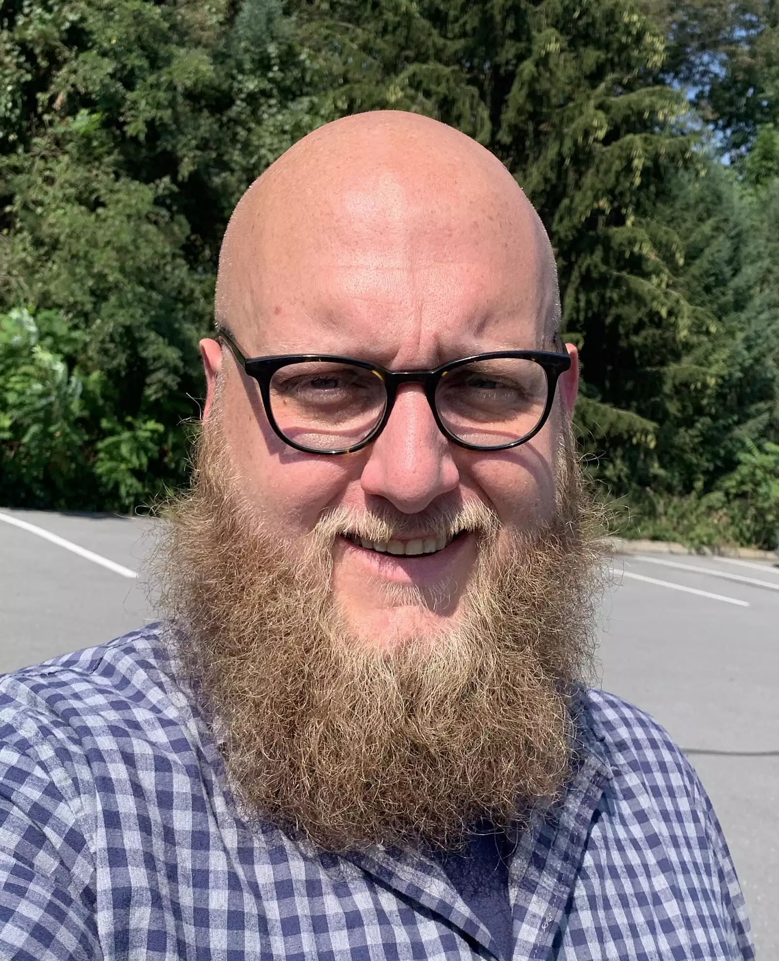 A smiling person with a bald head, glasses, and a full beard is wearing a checkered shirt. They are outdoors, standing in a parking area with trees in the background and a sunny sky.