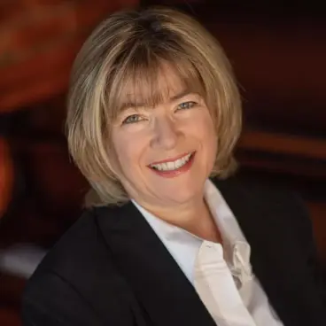 A person with short, light brown hair smiles warmly. They are wearing a white shirt and a black blazer. The background is softly blurred with warm tones.