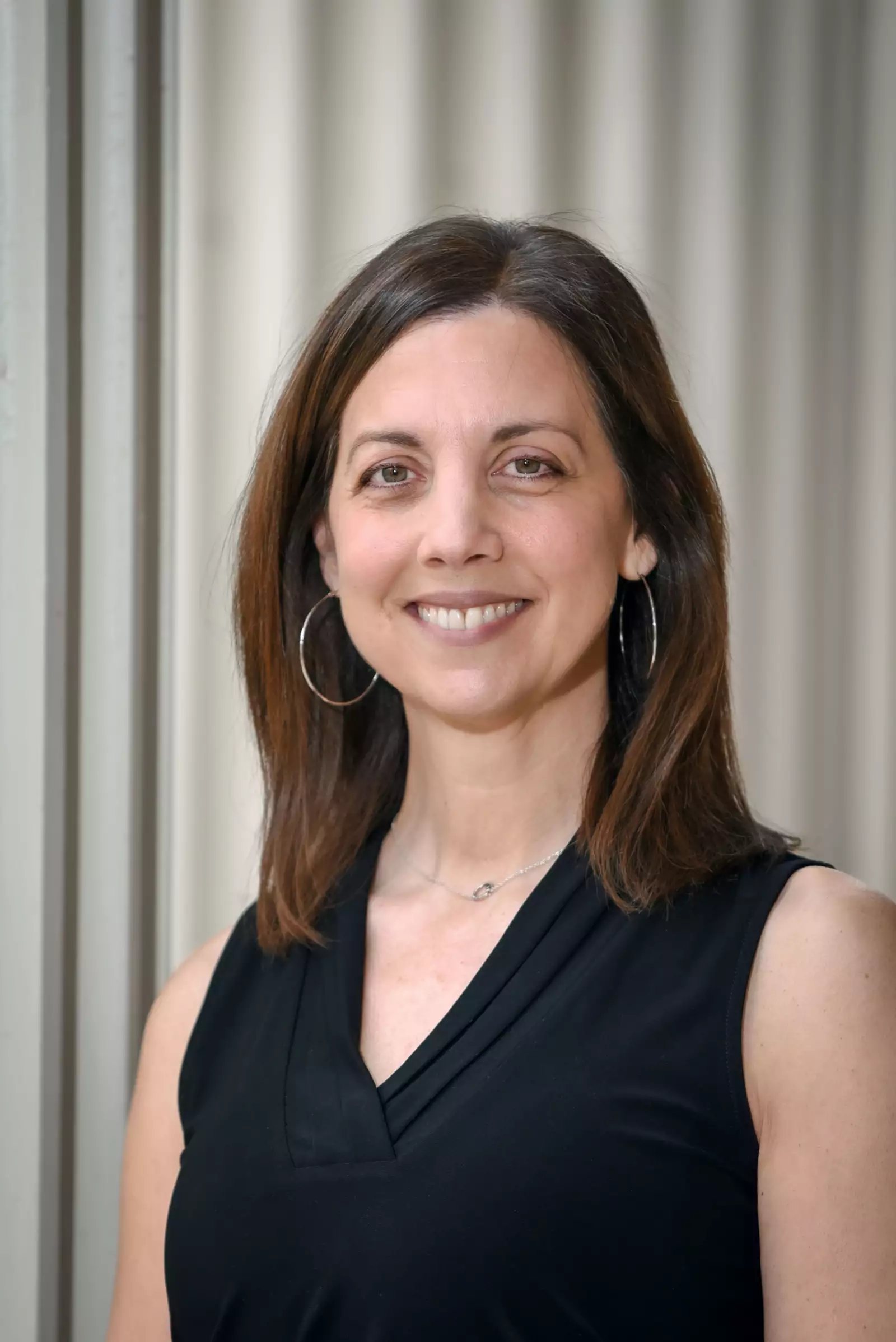 A woman with shoulder-length brown hair is smiling while standing in front of a neutral background. She is wearing a black sleeveless top and hoop earrings.