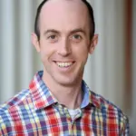 A person with short hair is smiling at the camera, wearing a plaid shirt with red, yellow, and blue stripes. The background is blurred, featuring vertical lines.