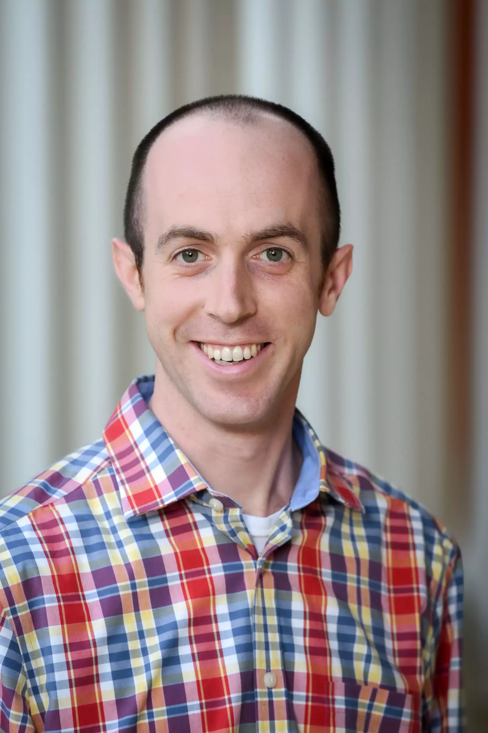 A person with short hair is smiling at the camera, wearing a plaid shirt with red, yellow, and blue stripes. The background is blurred, featuring vertical lines.