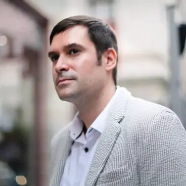A man in a light gray blazer and white shirt stands outdoors, gazing thoughtfully into the distance. The background is softly blurred, suggesting an urban environment.