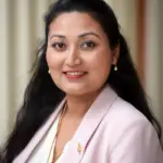A woman with long, dark hair is smiling. She is wearing a light pink blazer over a white blouse, accessorized with gold earrings and a necklace. The background features light-colored vertical stripes.