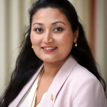 A woman with long, dark hair is smiling. She is wearing a light pink blazer over a white blouse, accessorized with gold earrings and a necklace. The background features light-colored vertical stripes.