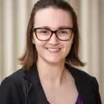 A person with shoulder-length brown hair and glasses smiles at the camera. They are wearing a black blazer over a purple top, standing against a neutral background.