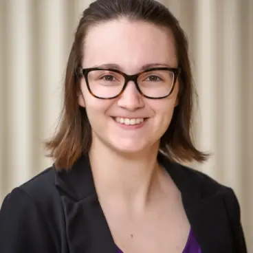 A person with shoulder-length brown hair and glasses smiles at the camera. They are wearing a black blazer over a purple top, standing against a neutral background.
