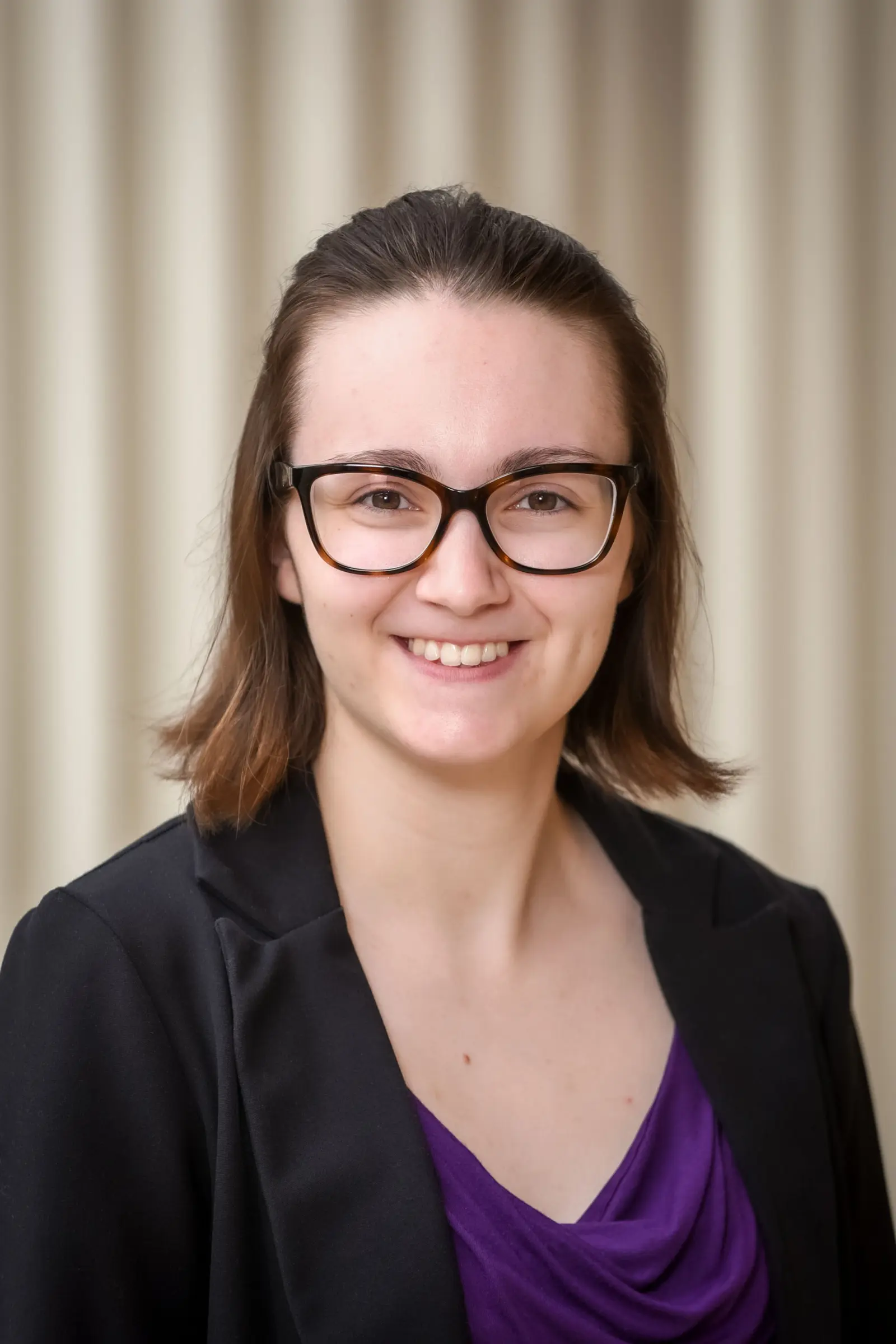 A person with shoulder-length brown hair and glasses smiles at the camera. They are wearing a black blazer over a purple top, standing against a neutral background.