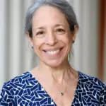 A woman with gray hair, smiling, wearing a blue patterned dress and a silver necklace. She is standing in front of a blurred light-colored background.