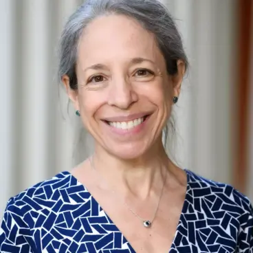 A woman with gray hair, smiling, wearing a blue patterned dress and a silver necklace. She is standing in front of a blurred light-colored background.