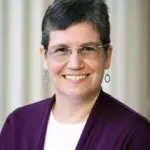 A person with short gray hair and glasses smiles at the camera. They are wearing a purple cardigan over a white shirt. The background features beige vertical stripes.