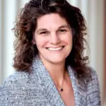 A woman with curly brown hair smiles while wearing a textured blazer over a dark top. She is standing in front of a blurred background of vertical light-colored lines, creating a warm and professional ambiance.