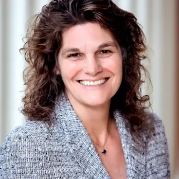 A woman with curly brown hair smiles while wearing a textured blazer over a dark top. She is standing in front of a blurred background of vertical light-colored lines, creating a warm and professional ambiance.