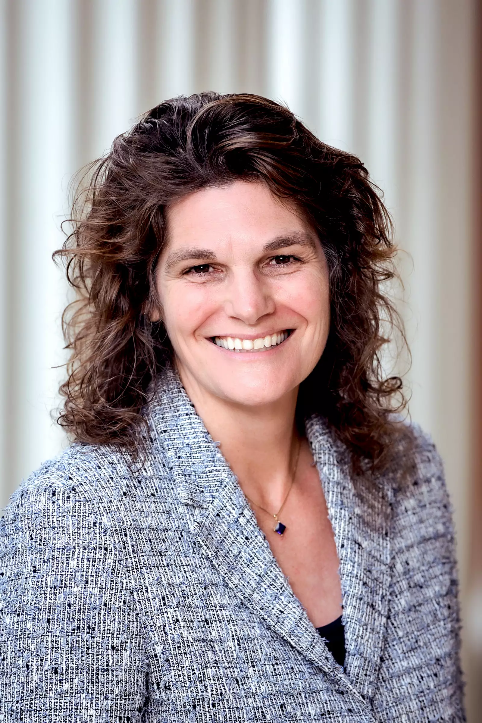 A woman with curly brown hair smiles while wearing a textured blazer over a dark top. She is standing in front of a blurred background of vertical light-colored lines, creating a warm and professional ambiance.