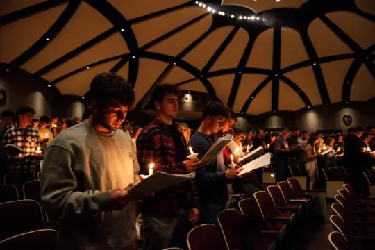 People participating in religious studies, standing in a dimly lit auditorium, each holding a lit candle and a program or song sheet. 