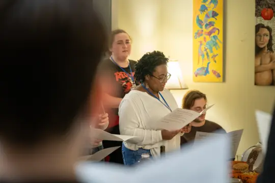 A diverse group of people standing indoors, each holding sheets of paper, possibly singing or rehearsing. Colorful artwork hangs on the wall behind them. The atmosphere appears focused and engaged.