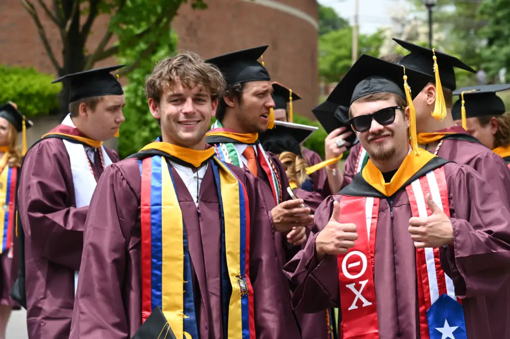 A group of graduates in maroon gowns and caps stand together outdoors. Two graduates in the foreground smile; one gives a thumbs-up. They wear colorful stoles and cords, and trees and a building are visible in the background.