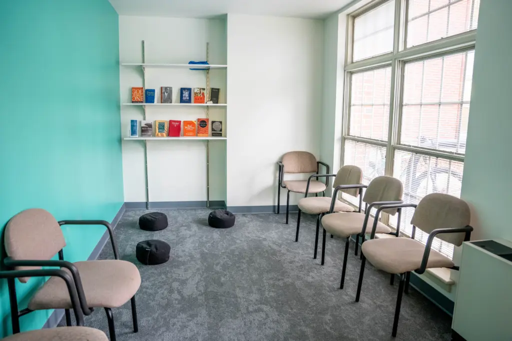 A small room with green and white walls features six beige chairs arranged in a semicircle on a carpeted floor. A shelf holds several books, and three round black cushions are placed on the floor. Large windows let in natural light.