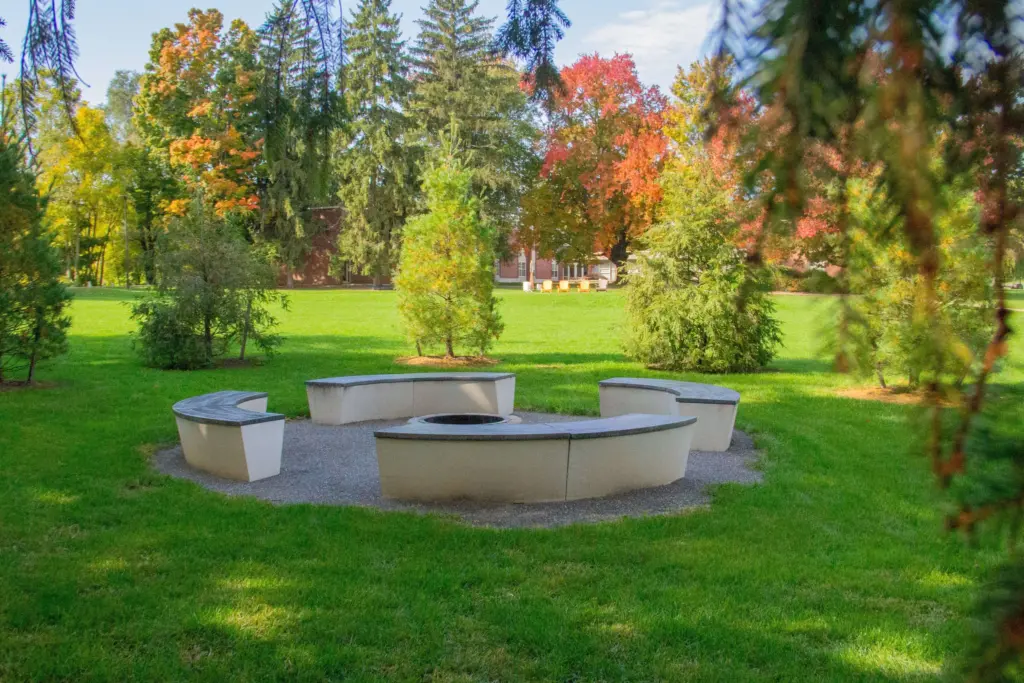 A circular stone fire pit surrounded by curved benches is set on a grassy field with tall trees, some showing autumn colors, in the background. The area is peaceful and vibrant under a clear blue sky.