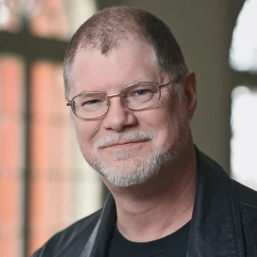 A man with short hair and glasses, wearing a black leather jacket over a black shirt, smiles softly. He has a trimmed beard and stands in front of a window with a soft, blurred background.