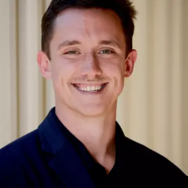 A person with short brown hair is smiling while wearing a dark blazer and a black shirt. The background is a light-colored wall with vertical lines.