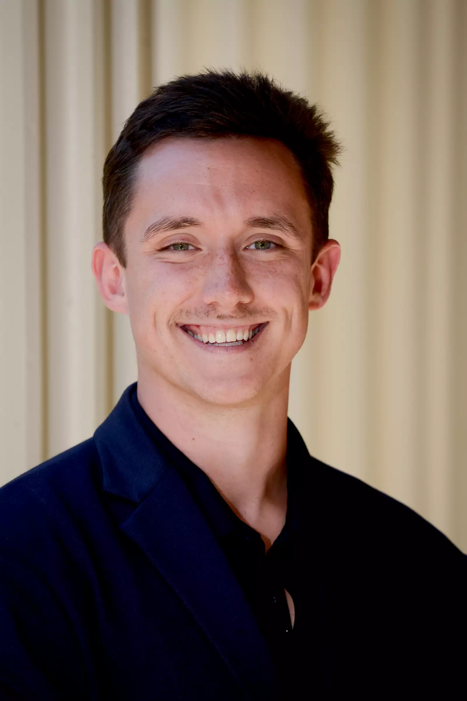 A person with short brown hair is smiling while wearing a dark blazer and a black shirt. The background is a light-colored wall with vertical lines.