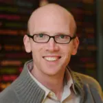 A person with glasses and a shaved head smiles at the camera. They are wearing a light-colored shirt under a gray sweater. The background is blurred with colorful text.