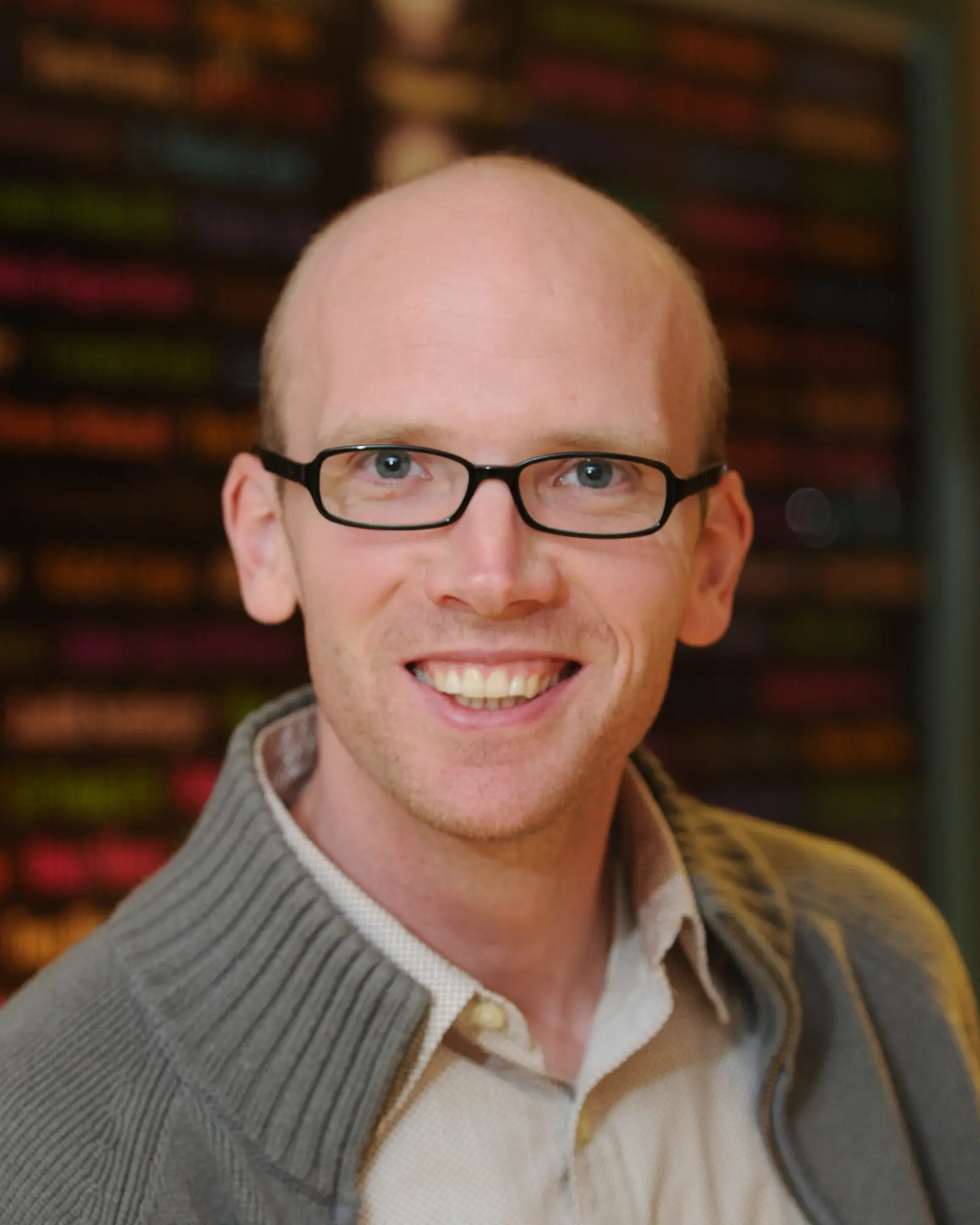 A person with glasses and a shaved head smiles at the camera. They are wearing a light-colored shirt under a gray sweater. The background is blurred with colorful text.