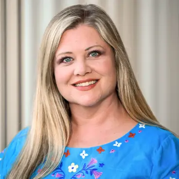 A woman with long blonde hair smiles at the camera. She is wearing a blue blouse with colorful floral embroidery and stands in front of a blurred, light-colored background.