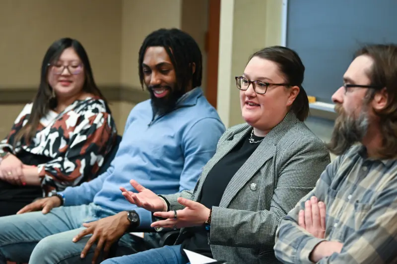 A group of four people sitting in a row, engaged in discussion. The woman with glasses in the center is speaking, gesturing with her hands. Others are attentively listening. They are in a room with a chalkboard behind them.