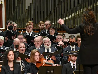 A choir of men and women in formal attire, many holding sheet music, sing while a conductor leads them. Some choir members are wearing masks. A string musician is visible in the foreground. The background features tall organ pipes.