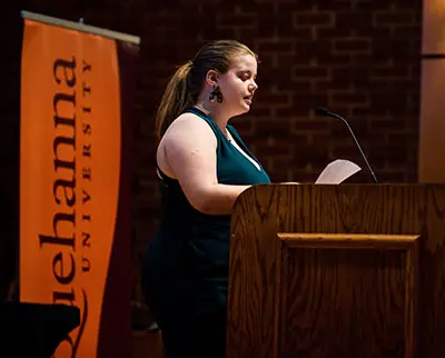 A person stands at a wooden podium, holding papers and speaking into a microphone. A banner with 