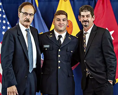 Three men are standing together in front of flags. The man in the middle is wearing a military uniform, while the men on either side are in suits. They are smiling and posing for a photo.