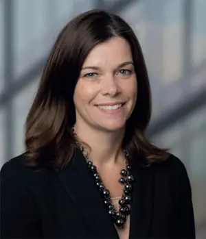 A woman with shoulder-length brown hair is smiling at the camera. She is wearing a black blazer and a pearl necklace, standing in front of a blurred background.
