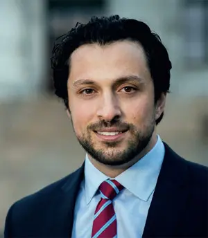 A man with dark hair and a beard, wearing a dark suit, light blue shirt, and red striped tie, is smiling and facing the camera. The background is blurred and neutral.