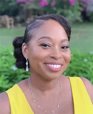 A person with a styled updo and earrings smiles warmly. They are wearing a yellow top and a delicate necklace. The background features greenery and pink flowers, suggesting an outdoor setting.