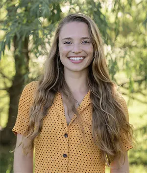 A woman with long, wavy hair smiles while standing outdoors. She is wearing a yellow polka dot blouse, and there are green trees softly blurred in the background, suggesting a sunny day.