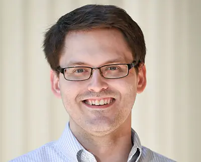 A person with short dark hair and glasses is smiling at the camera against a plain background, wearing a light-colored button-up shirt.