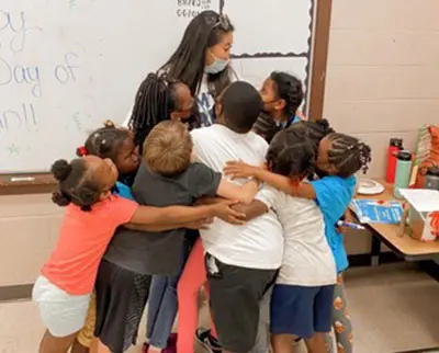 A group of children are gathered in a classroom, hugging a teacher. The children are smiling and appear happy. The teacher, wearing a face mask, stands by a whiteboard filled with writing. A table with various items sits nearby.