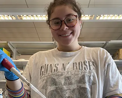 A person in a laboratory setting wearing a Guns N' Roses t-shirt and gloves is smiling while using a pipette. They have glasses and are standing under fluorescent lights with shelves in the background.