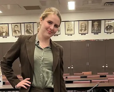 A person with blonde hair wearing a brown suit and green shirt stands confidently in a room. The background features framed black and white portraits on the wall.